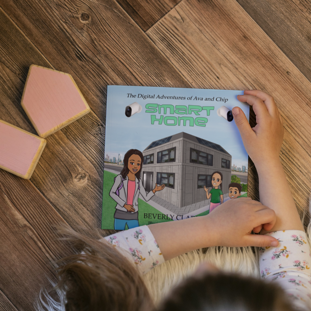 A child's hands hold open "The Digital Adventures of Ava and Chip: Smart Home" book, featuring a modern house on the cover, against a wooden floor with light pink wooden house-shaped blocks.